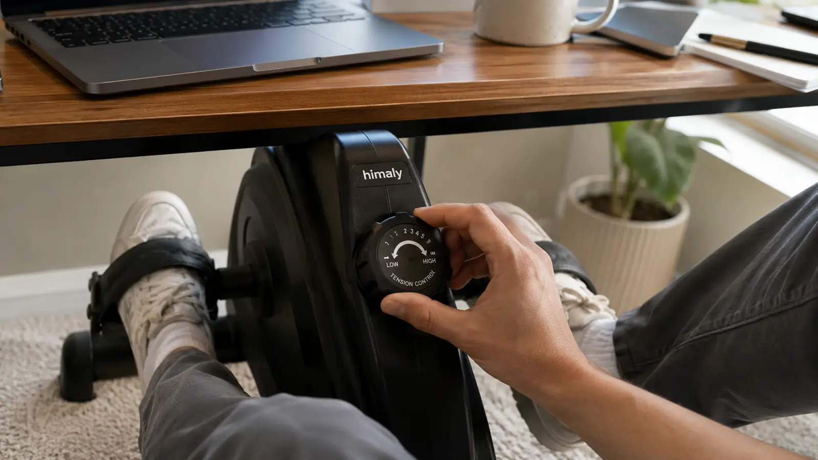 Hand adjusting the resistance dial on the himaly under-desk pedal exerciser while working at a laptop