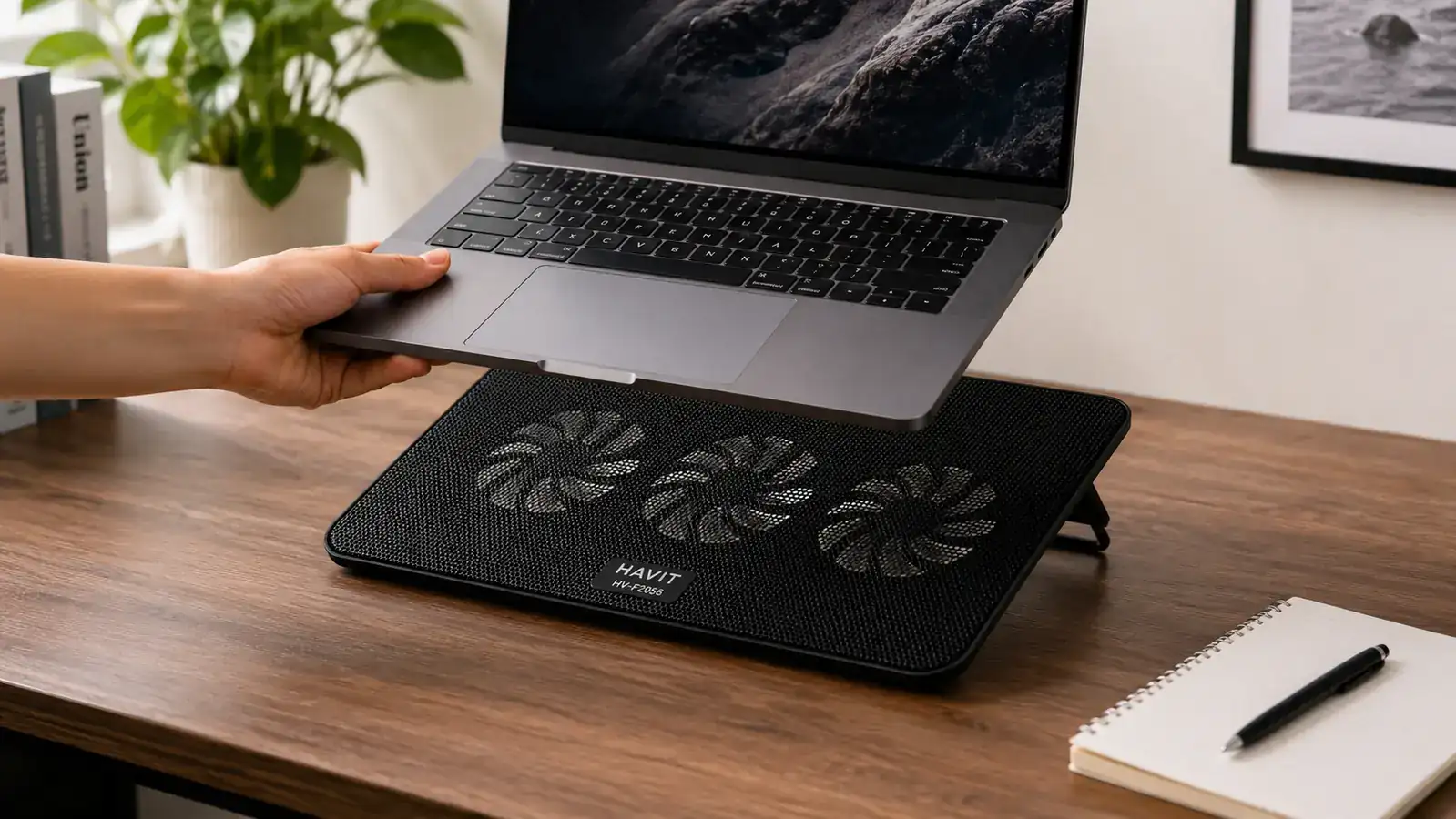 A hand placing a slim black laptop cooling pad with visible fan grilles under an open laptop on a desk