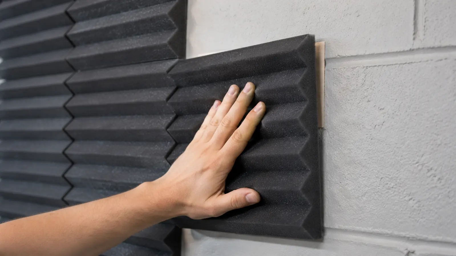 Hand pressing a foam acoustic panel corner against a painted cinderblock wall to test the adhesive strip